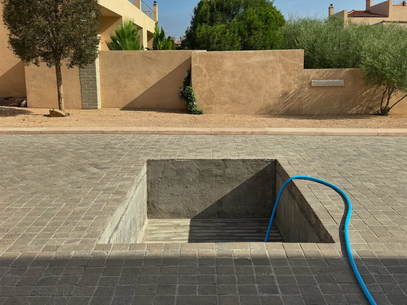 Open concrete underground water tank hatch in a villa driveway with a pump hose running out